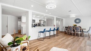 Ett modernt kök med vita skåp och en köksö, en matplats med blå stolar och ett träbord samt ett vardagsrum med en vit soffa och ett soffbord.