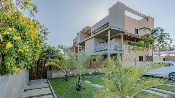 A modern two-story building with a balcony, surrounded by a well-maintained garden with palm trees and yellow flowering plants. - Houses in a Gated Community 100m From Marceneiro Beach - Hvas (Passo de Camaragibe)