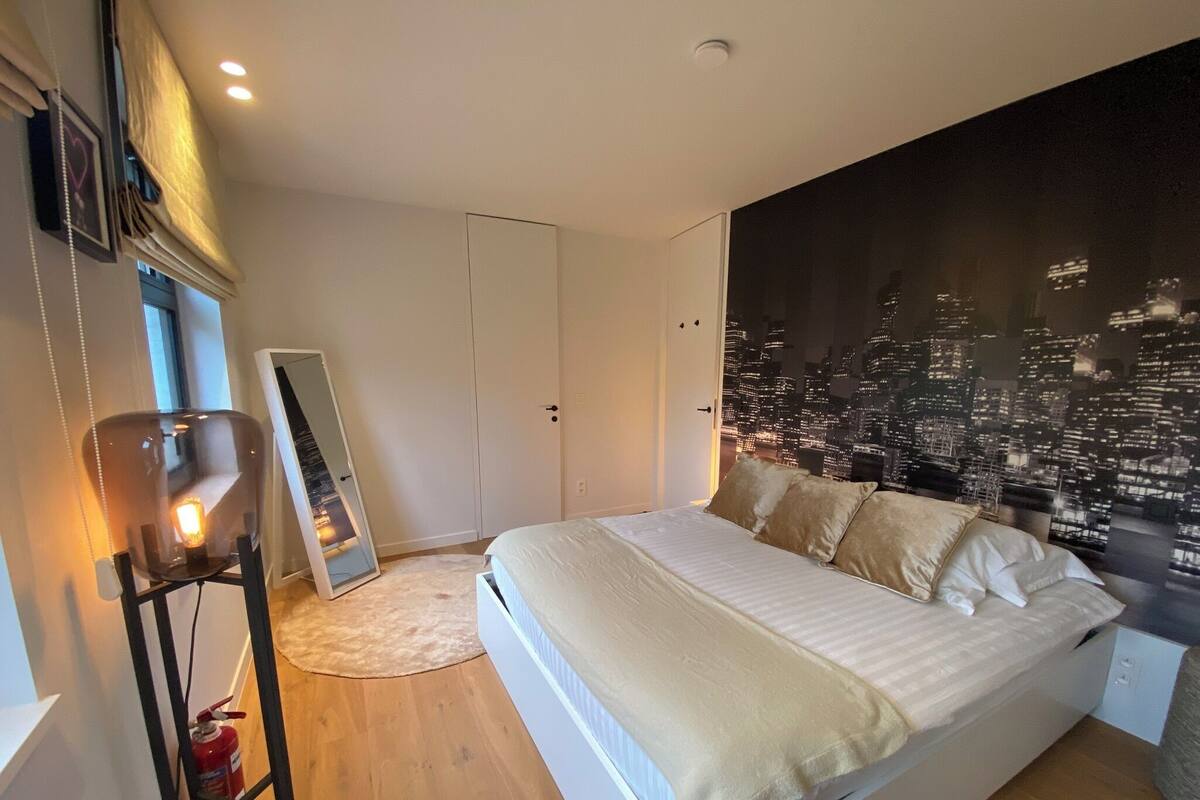 A bedroom with a cityscape mural, a bed with white linens, a bedside table, and a mirror.