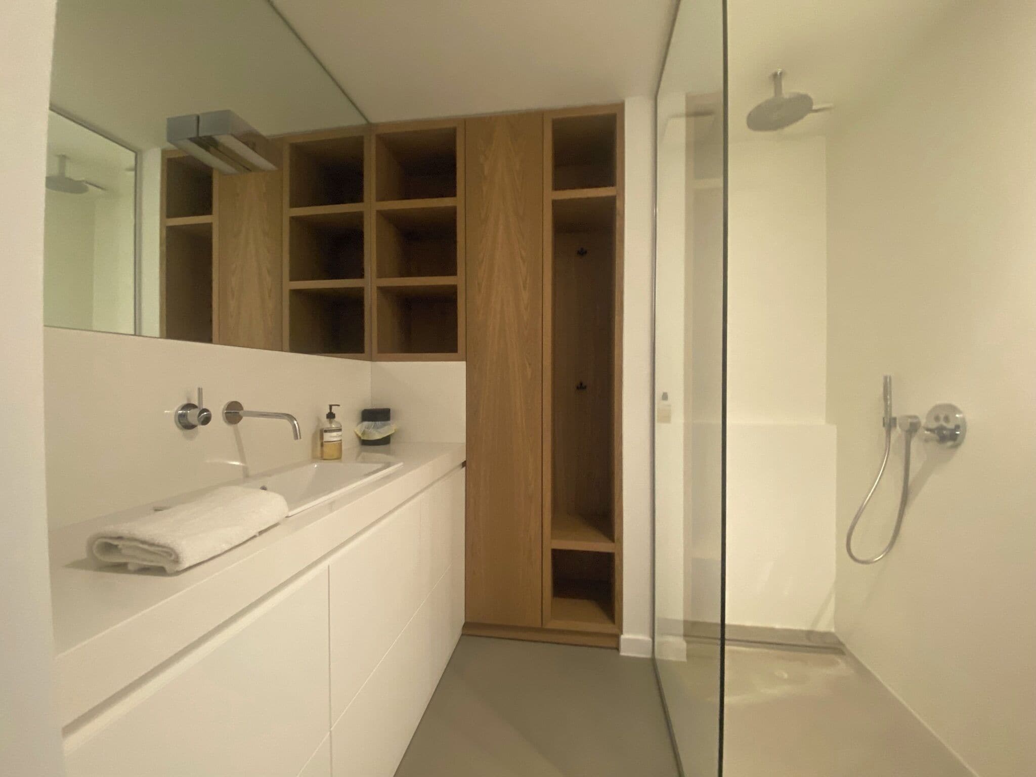 Un baño moderno con cabina de ducha de vidrio, tocador blanco con una toalla y estanterías de madera sobre el lavabo.
