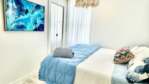 Un dormitorio con cama, lámpara colgante, un cuadro y ventana con cortinas.