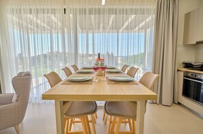 A dining area with a wooden table set for six, surrounded by chairs, with a view of the sea through sheer curtains. - Arilllas Villas Marilou (Corfu)