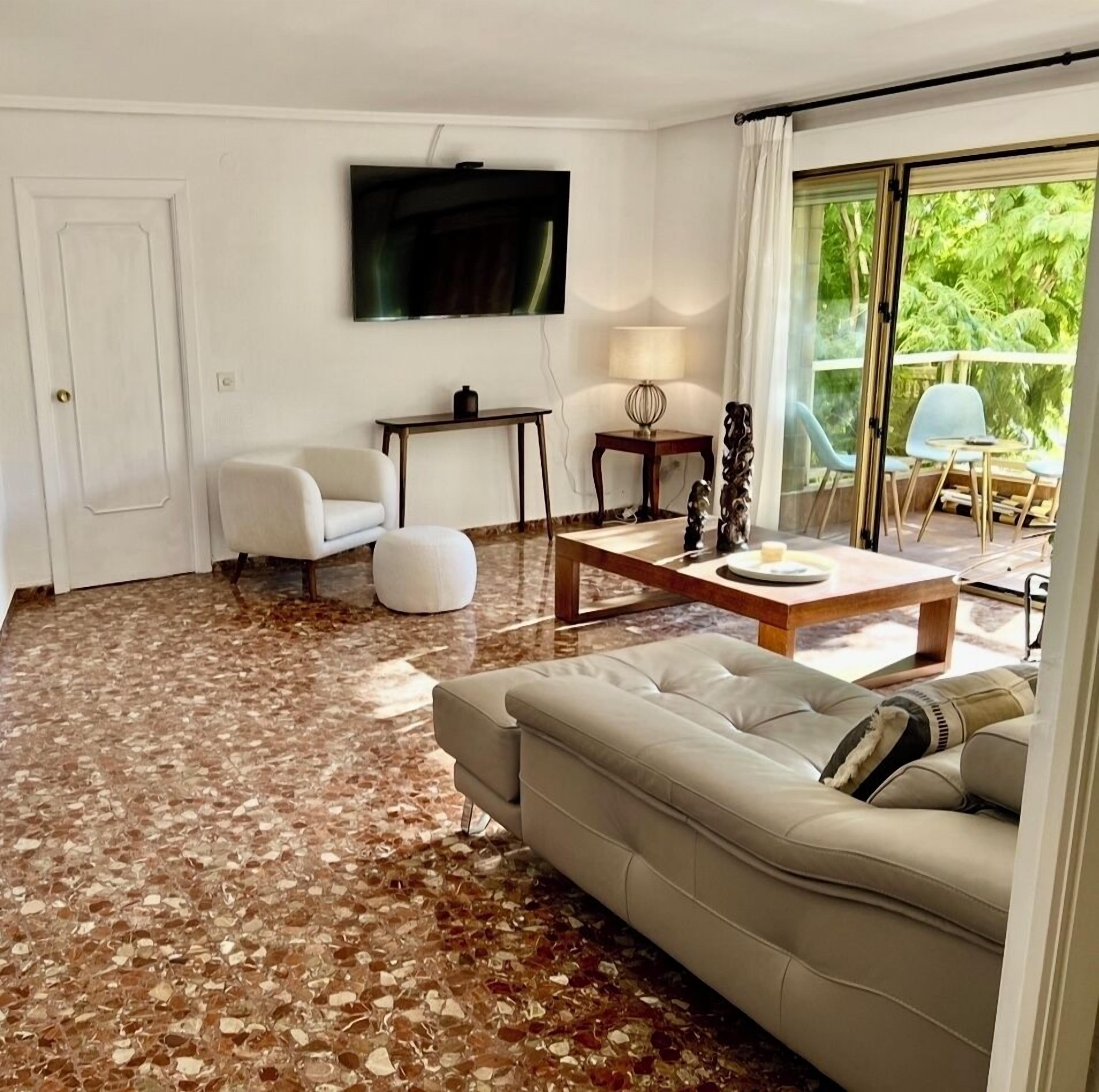Uma sala de estar com sofá, mesa de centro, TV fixada na parede e vista para o verde pela janela.