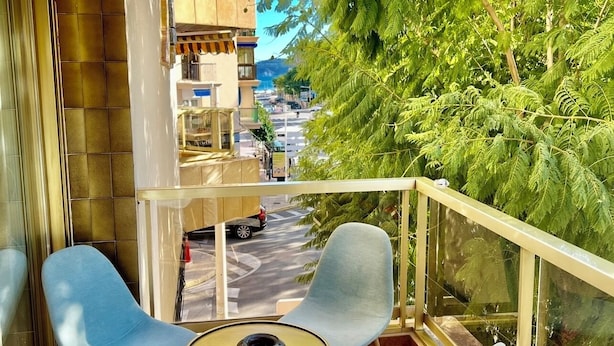 Un balcon avec deux chaises et une petite table, donnant sur une rue et une végétation luxuriante.