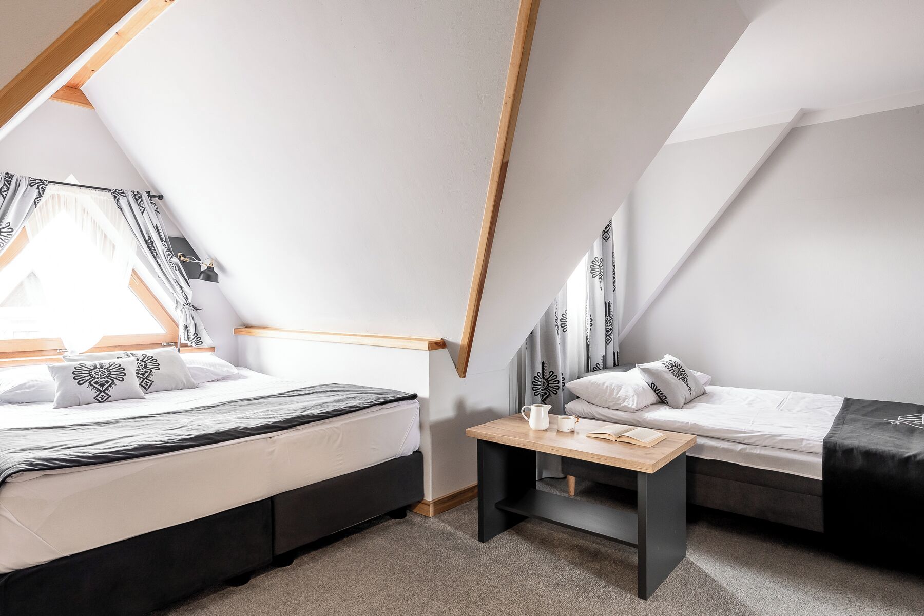 A bedroom with a sloped ceiling, two beds, a small table, and a window with curtains.
