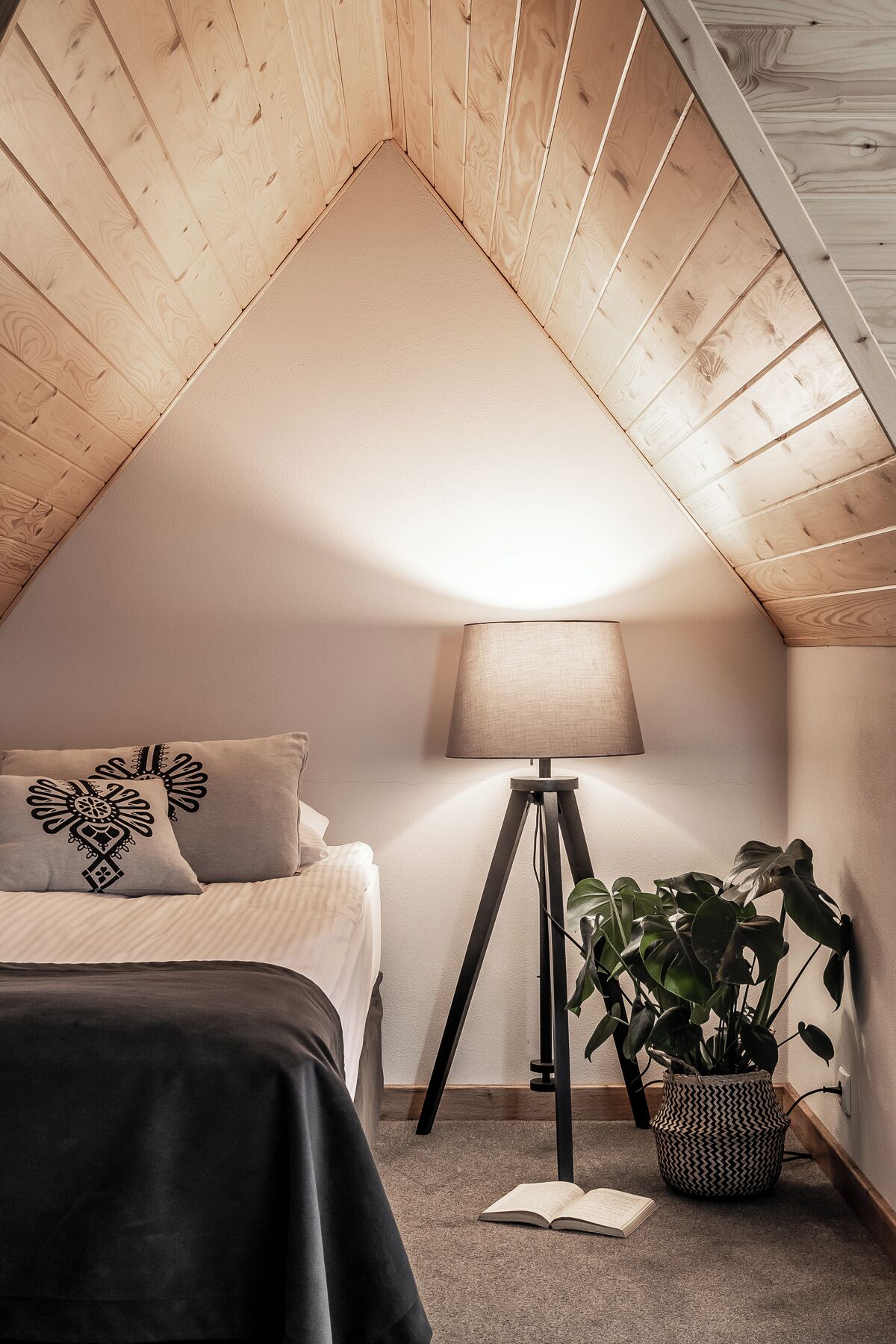 A cozy attic bedroom with a sloped ceiling, a bed with patterned pillows, a bedside lamp, a potted plant, and an open book on the floor.