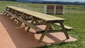 A row of wooden picnic tables with benches, set on a paved area with a grassy field and a fence in the background. - Group and stopover gîte in the heart of nature, on the Chemin de St Jacques GR65-GR7 (Riotord)