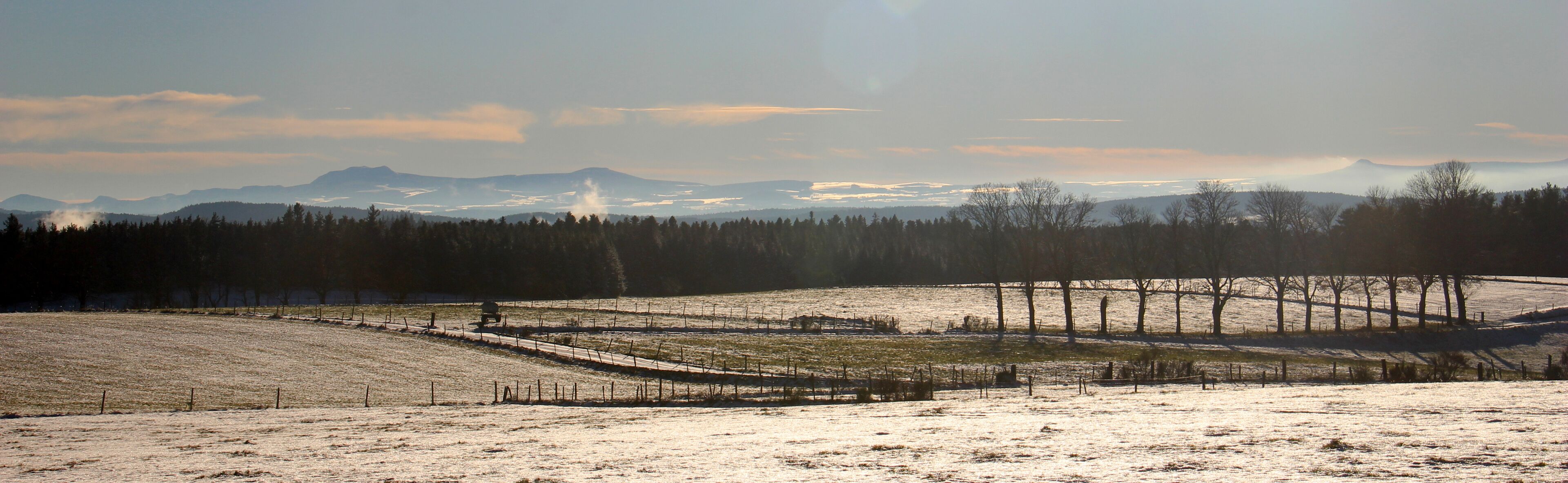 Een besneeuwd landschap met een hek en bomen, en verre bergen onder een heldere lucht.
