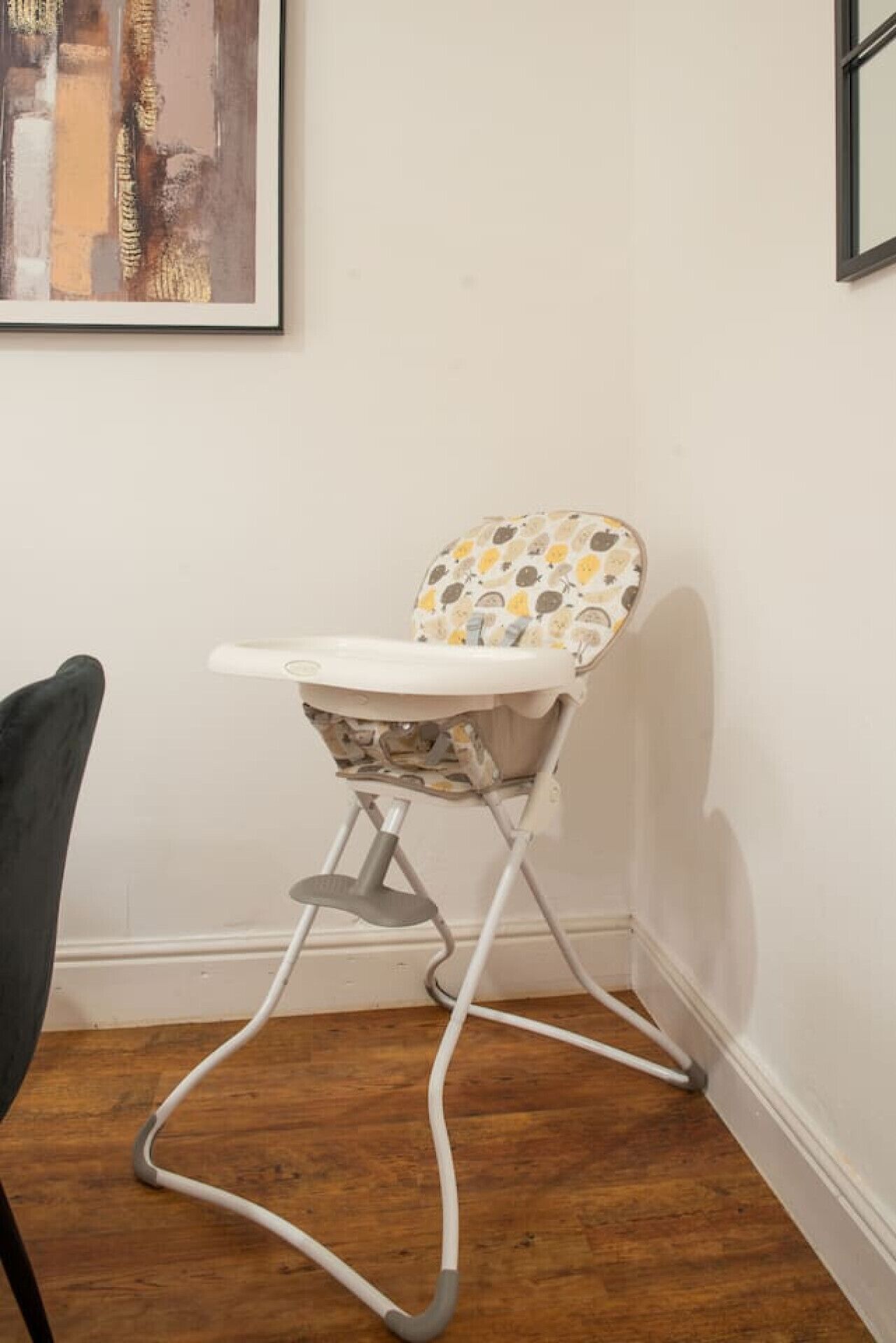 En babystol med et mønstret sæde og en hvid bakke, placeret op ad en hvid væg med et billede over.
