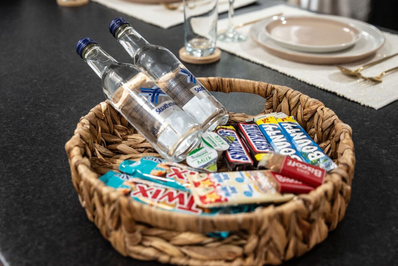 En korg av rotting som innehåller flaskor, snacks och ett glas, placerad på ett bord.