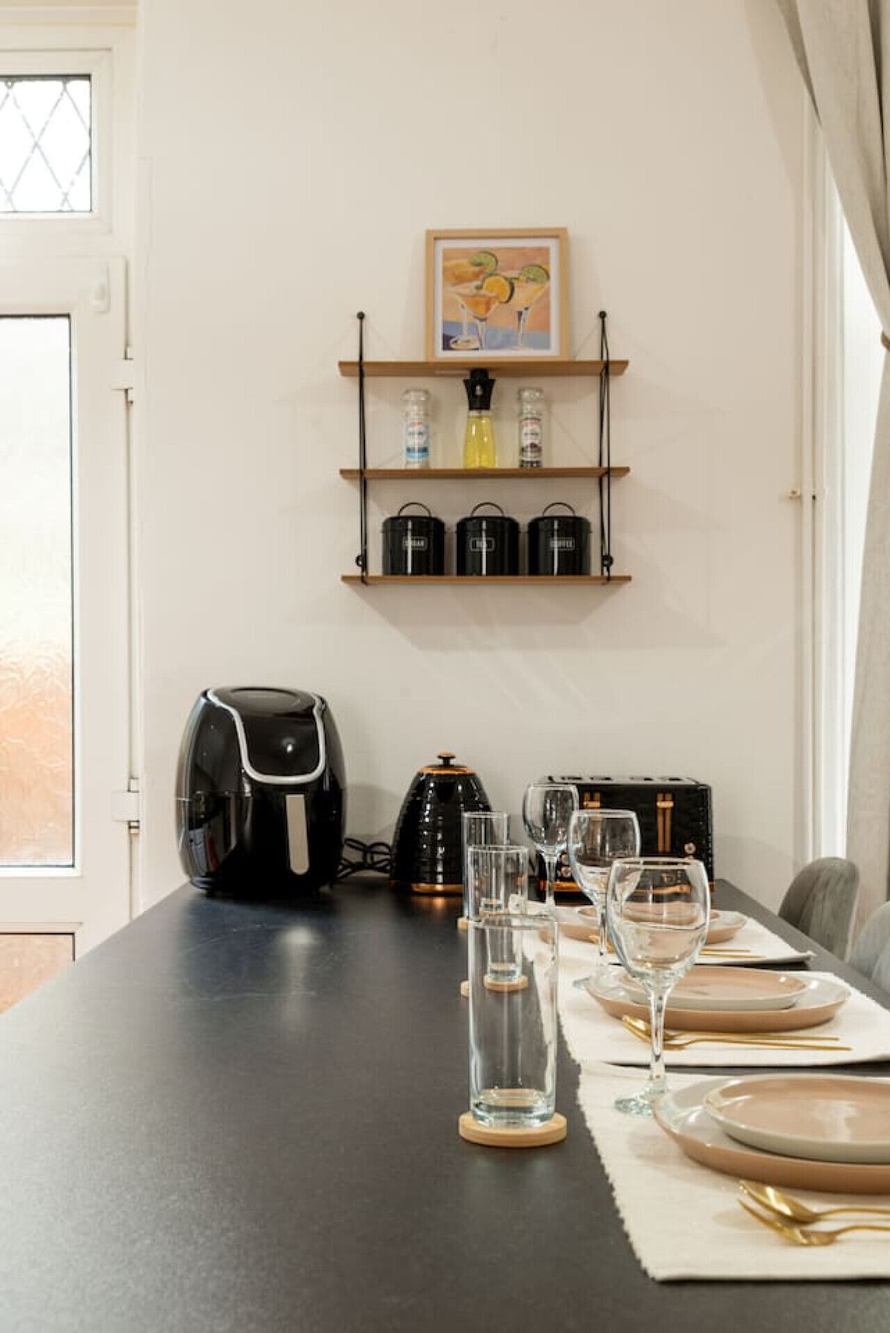 Una mesa de comedor dispuesta con platos, vasos, cubiertos, una cafetera y un hervidor. Encima de la mesa hay estantes con frascos y un cuadro enmarcado.