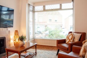 Un salon avec un canapé, une table basse, un téléviseur et une grande fenêtre équipée de stores.