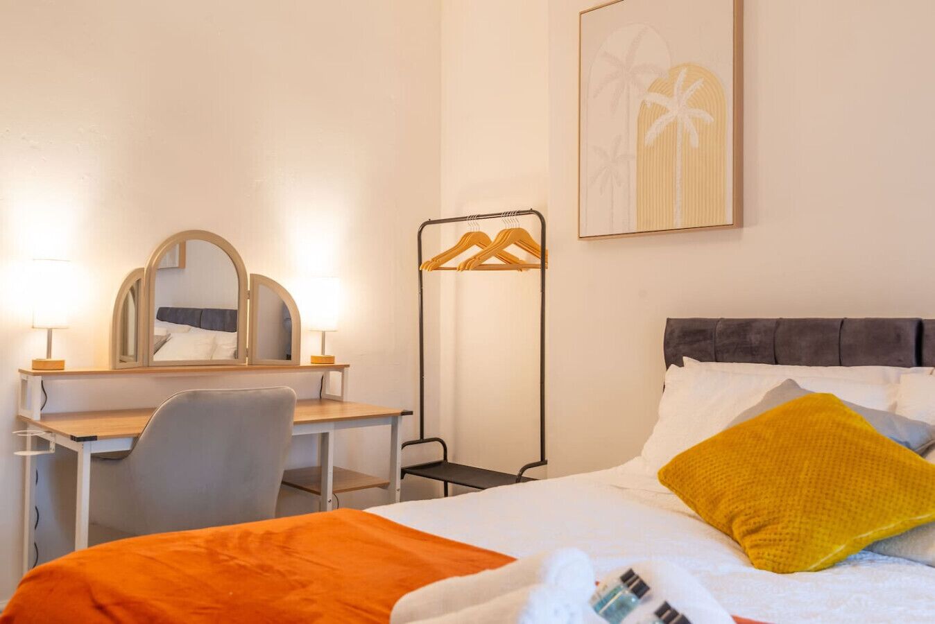 Ein Hotelzimmer mit einem Bett, einem Schreibtisch mit Stuhl, einem Spiegel, einem Nachttisch, einer Lampe und einem gerahmten Bild an der Wand.