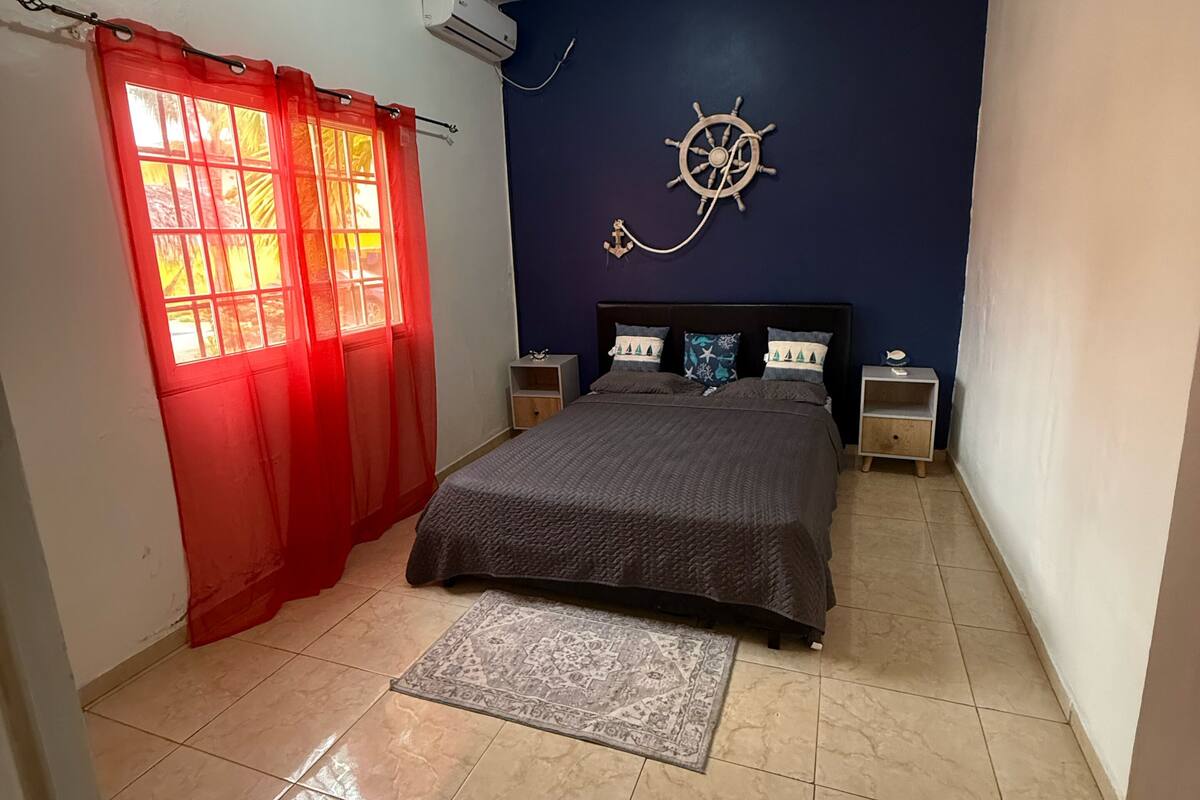 Een slaapkamer met een bed, nachtkastjes, een aan de muur bevestigd stuurwiel, een rood gordijn en een betegelde vloer.