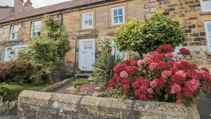 A stone building with a red-tiled roof, a white door, and a small garden with red flowers and green shrubs. - Manor Cottage (Scarborough)