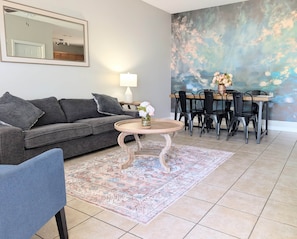 A living room with a grey sofa, a round coffee table, a blue armchair, a dining table with chairs, and a large abstract painting on the wall. - Alabama Daydreaming 3BD/2.5 bath *5 min to base*pool (Enterprise)