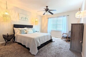 Un dormitorio con cama, ventilador de techo, ventana con persianas, una silla y una lámpara decorativa.