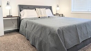 Une chambre à coucher comprenant un lit, deux tables de chevet, une commode, une lampe et une fenêtre avec des stores.