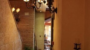 A hallway with hexagonal tiled flooring, a wooden ceiling, and a brick wall on one side. There are hanging lamps and a potted plant on the left. - Hotel Doña Isabel (Elche)