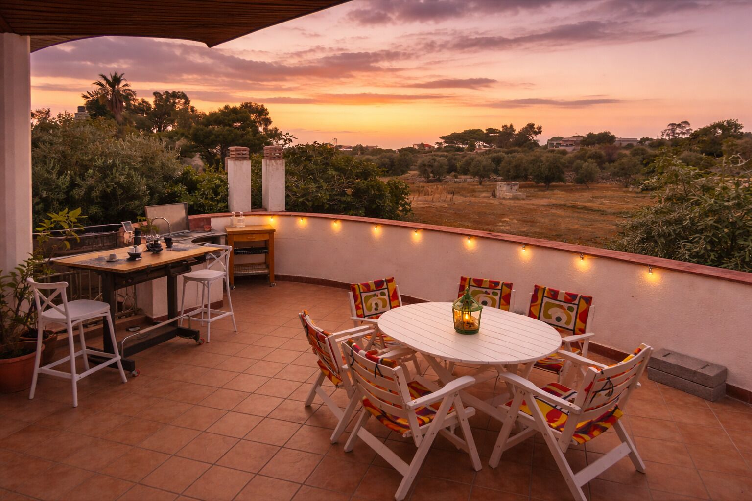 Un espacio al aire libre cubierto con mesa y sillas, rodeado de plantas y vistas al paisaje.