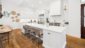 A modern kitchen with white cabinetry, a large island, and a dining area with wooden flooring and metal chairs. - Gulf Getaway with Private Pool and LSV (Santa Rosa Beach)