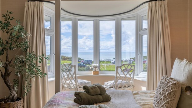 Ein Schlafzimmer mit einem großen Erkerfenster, einem Bett mit aufgerollten Handtüchern, einer Topfpflanze und Blick auf das Meer und geparkte Autos.