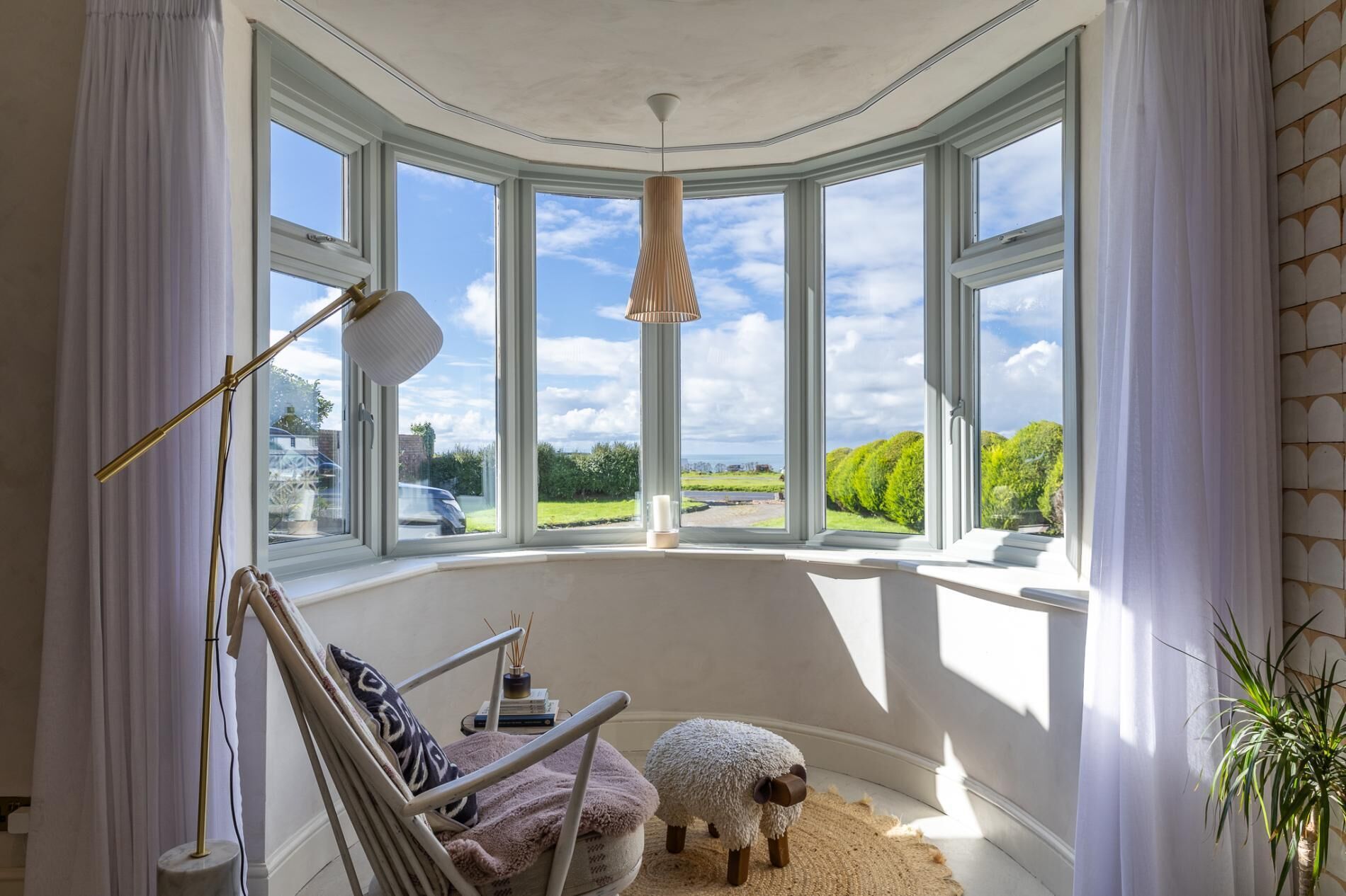 Ein gemütliches Zimmer mit einer gewölbten Erkerfensterfront, einem bequemen Sessel, einem kleinen Tisch mit Kerze und einer dekorativen Schaf-Figur.