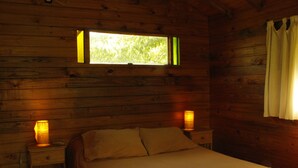 Ein Zimmer in einer Holzhütte mit einem Bett, zwei Nachttischlampen und einem Fenster mit Blick auf Grün.