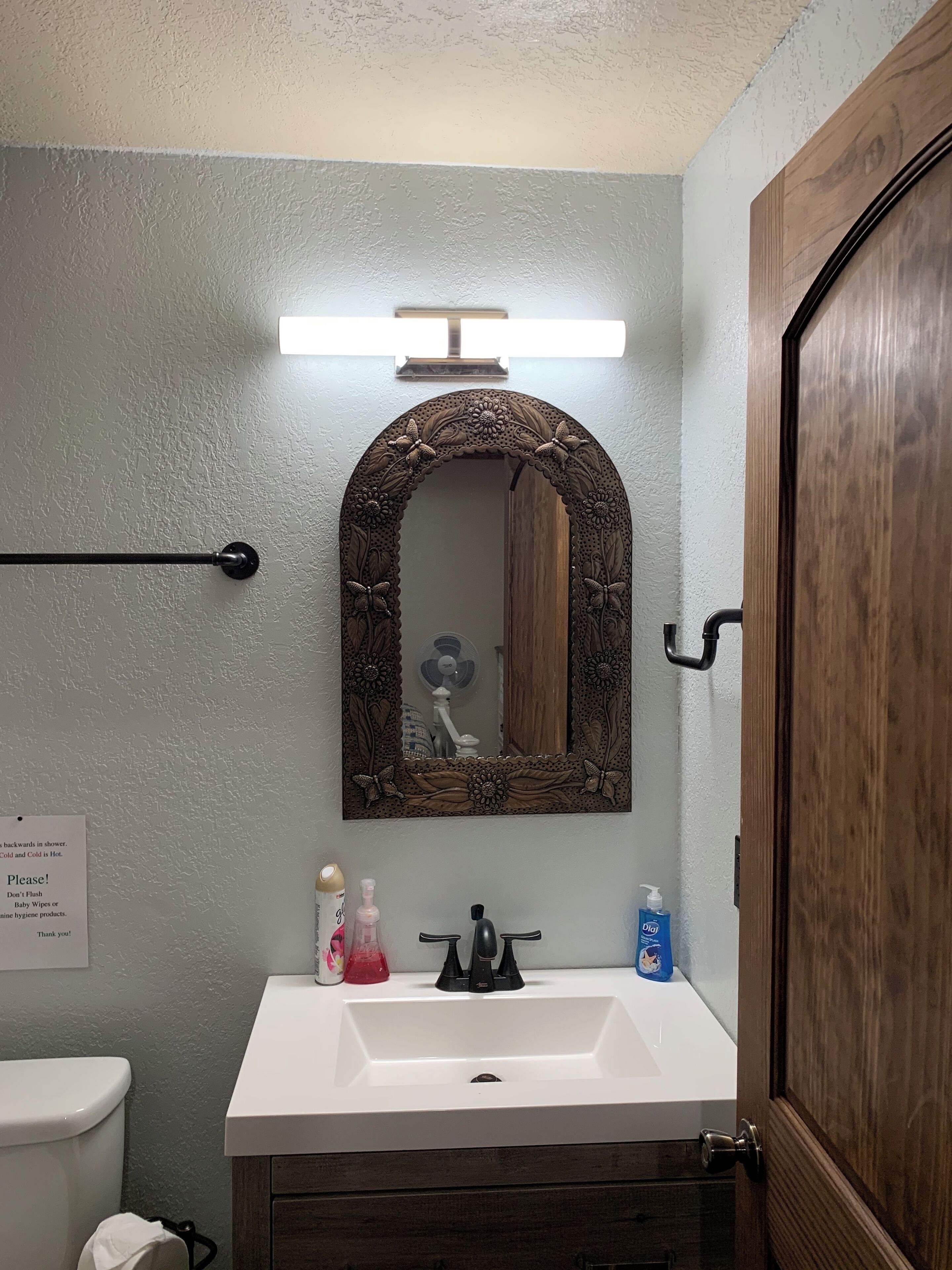 Une salle de bain avec un lavabo blanc, une porte en bois, un miroir orné de motifs floraux et un porte-serviettes.