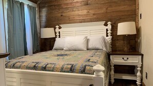 A bedroom with a wooden ceiling, a white bed with a colorful quilt, two bedside lamps, and a nightstand with a drawer. - Rustic Cabin Retreat Near the Nueces River (Camp Wood)