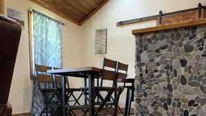 A dining area with a stone accent wall, wooden ceiling beams, and a rustic wooden table and chairs. - Rustic Cabin Retreat Near the Nueces River (Camp Wood)