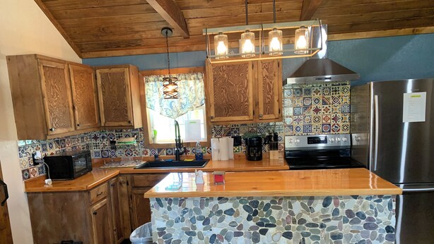 A kitchen with wooden cabinets, a tiled backsplash, a central island with stools, a stainless steel refrigerator, and a hanging light fixture. - Rustic Cabin Retreat Near the Nueces River (Camp Wood)