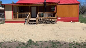 A single-story building with a red roof and wooden siding, featuring a front porch with a wooden railing and a set of stairs leading to the entrance. - Rustic Cabin Retreat Near the Nueces River (Camp Wood)