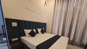 Habitación de hotel con cama, cabecero, mesita de noche, lámpara y ventana con cortinas.