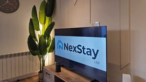 Un salon avec un téléviseur à écran plat affichant le logo de « NexStay Ltd », une plante en pot, un radiateur et une horloge murale.