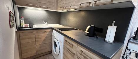 Een compacte keuken met houten kastjes, een wasmachine, een spoelbak en een koffiezetapparaat.