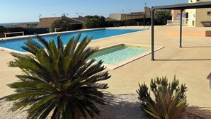 Piscina com vista para o mar, cercada por palmeiras e um edifício com telhado de azulejos.