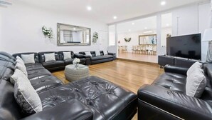 Un salon moderne avec un canapé d’angle, un téléviseur à écran plat et un coin repas visible à travers une porte ouverte.