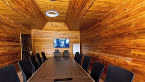 A conference room with a long wooden table, black chairs, a whiteboard, and a mounted screen.