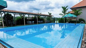 A swimming pool with tiled edges, a covered seating area with chairs, and a building with a tiled roof in the background.