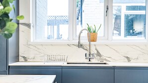 Une cuisine dotée d’un plan de travail en marbre, d’un évier en acier inoxydable et d’une fenêtre donnant sur une maison et une clôture.