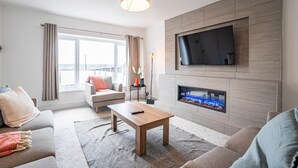 Un salon moderne avec un téléviseur à écran plat fixé à un mur en bois, une cheminée, un canapé, une table basse et une grande fenêtre avec des rideaux.