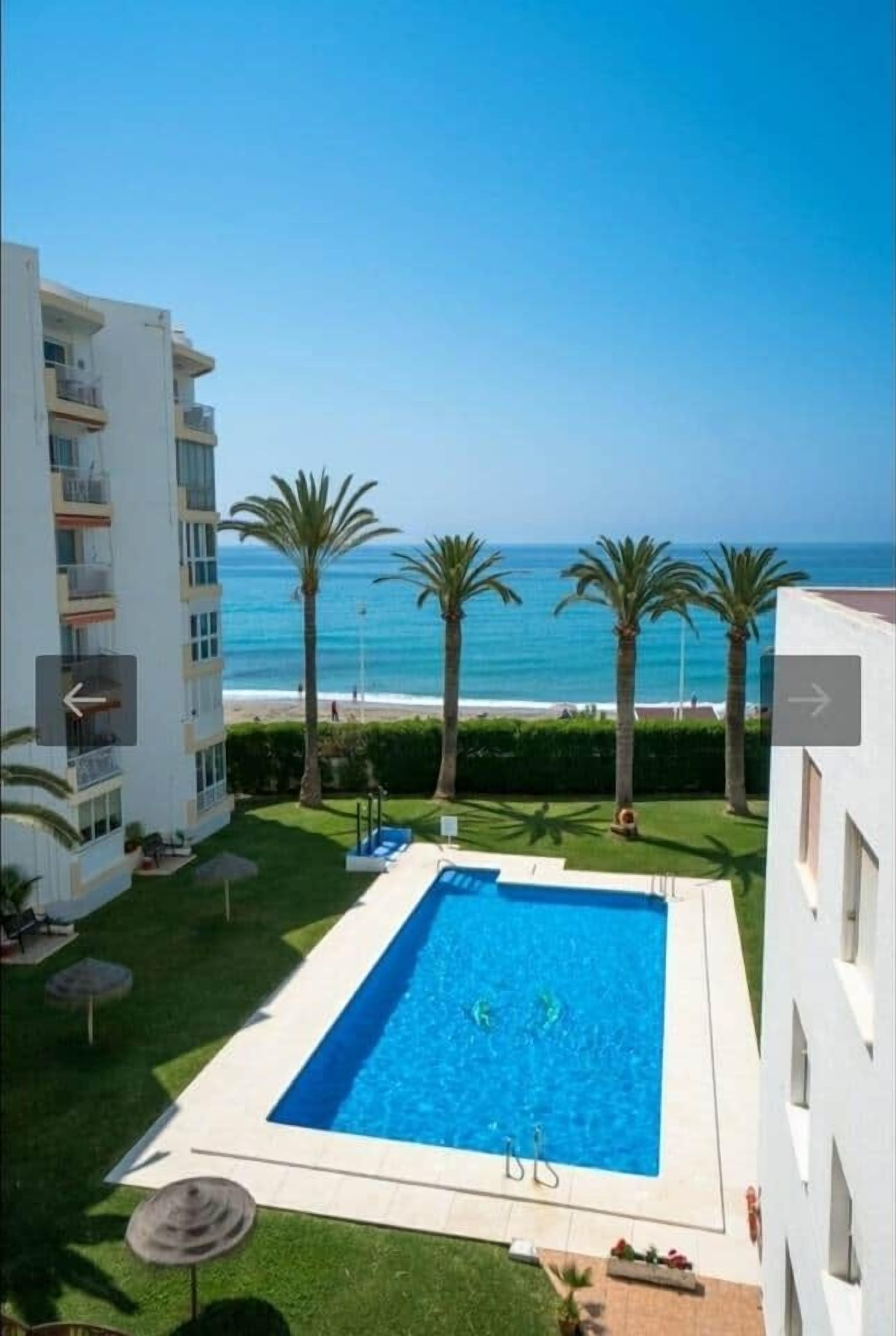 Piscina com vista para o oceano, cercada por palmeiras e um edifício.