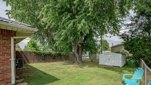 A backyard with a large tree, a brick house, and two Adirondack chairs.