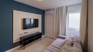 A modern living room with a flat-screen TV mounted on a white wall, a black entertainment unit, a light-colored sofa, and a window with curtains. - Casa em Jurerê com 4 quartos - 10 min da praia (Florianópolis)
