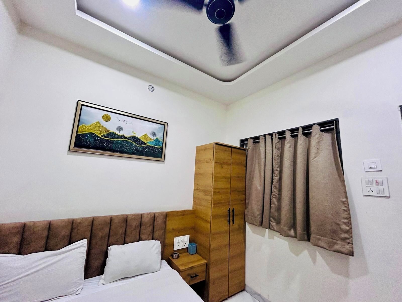 A hotel room with a bed, a wooden wardrobe, a nightstand, a power outlet, and a ceiling fan.