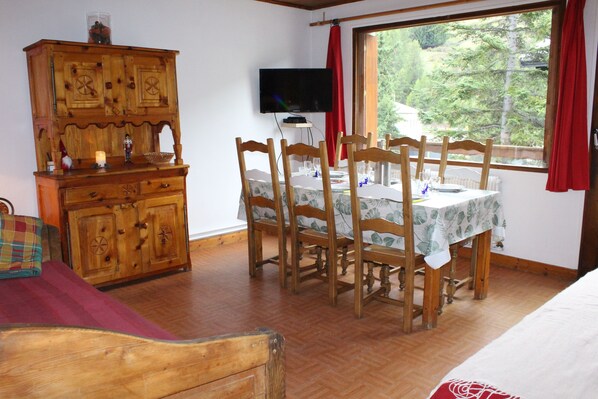 Ein Zimmer mit einem Holzesstisch, Stühlen, einem Fernseher und einem Fenster mit Blick auf Bäume.