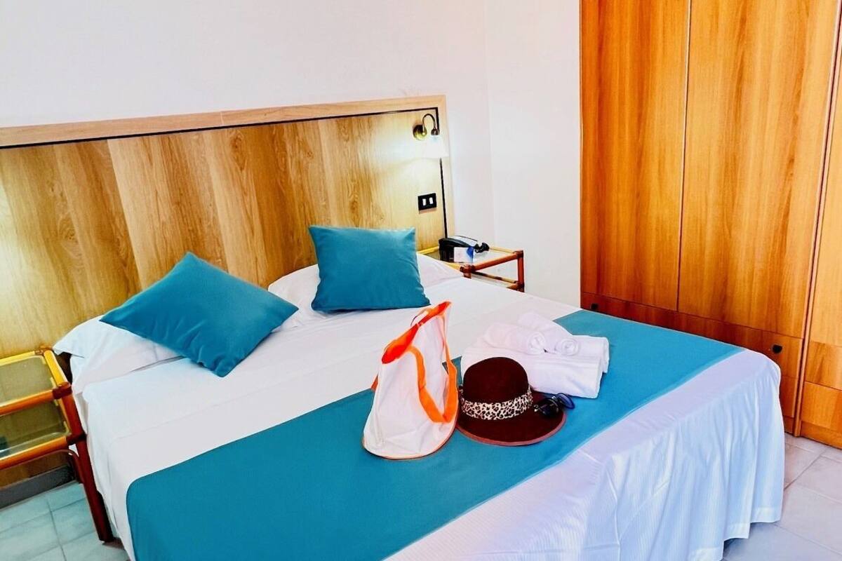 A hotel room with a large bed, a wooden wardrobe, and a small bedside table.