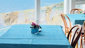 Une table recouverte d’une nappe bleue, des chaises et une vue sur la mer par de grandes fenêtres.