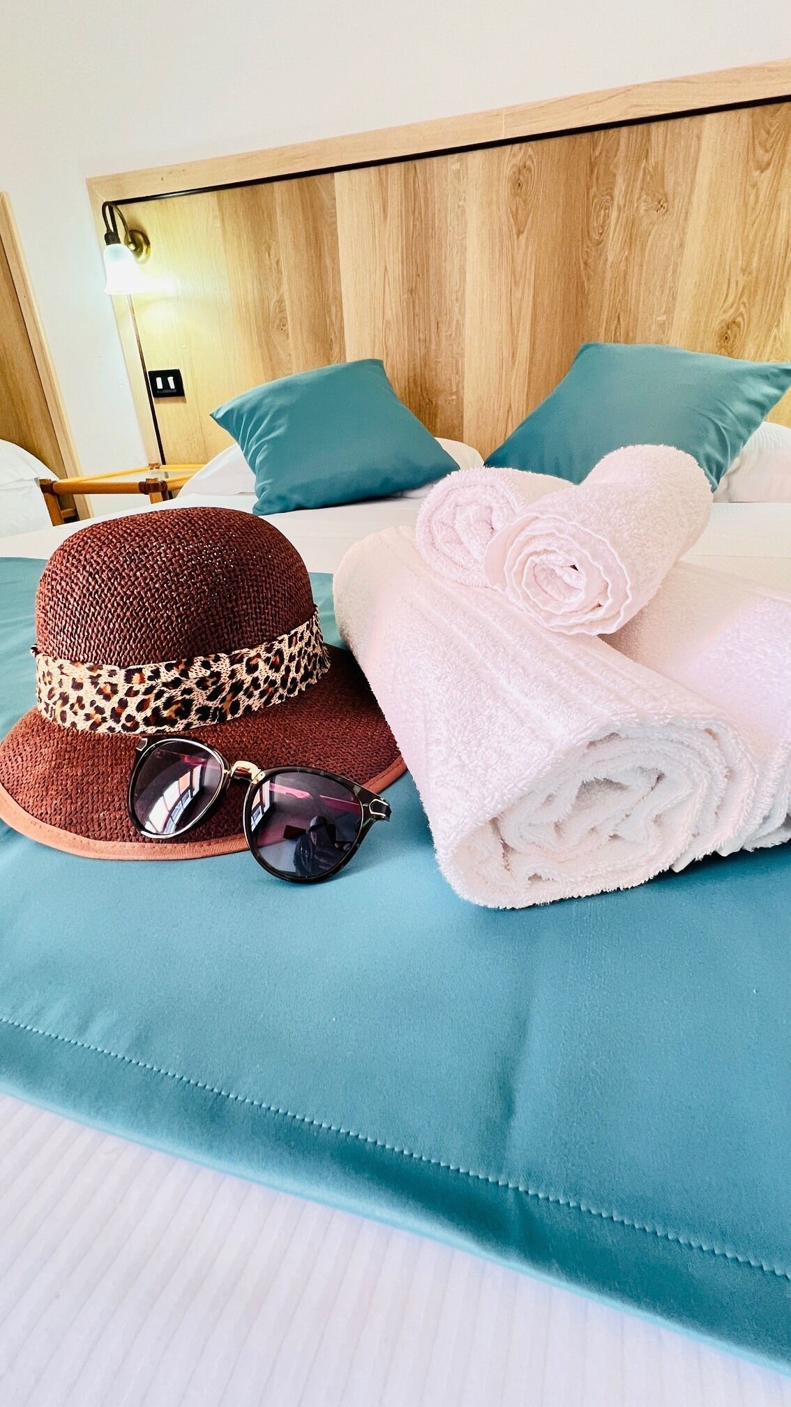 Ein ordentlich bezogenes Bett mit türkisfarbenem Bettwäsche, ein brauner gewebter Hut mit Leopardenmuster-Band, Sonnenbrille und aufgerollte Handtücher.